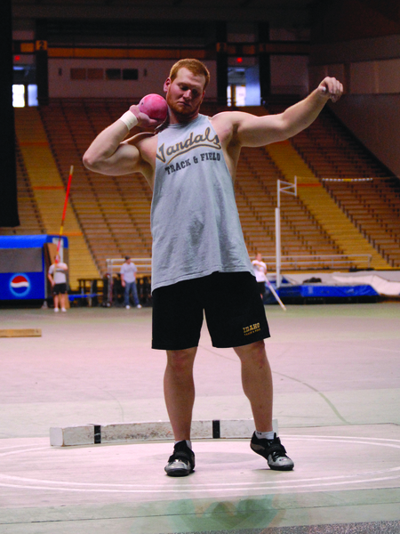 Color photograph of a man holding a shot put by his neck. In the background are empty stadium stands.